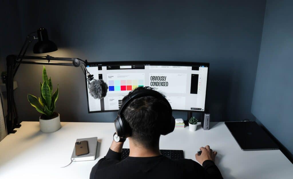A person with headphones is working at a desk with a large monitor displaying a design interface, surrounded by a plant, lamp, and gadgets.