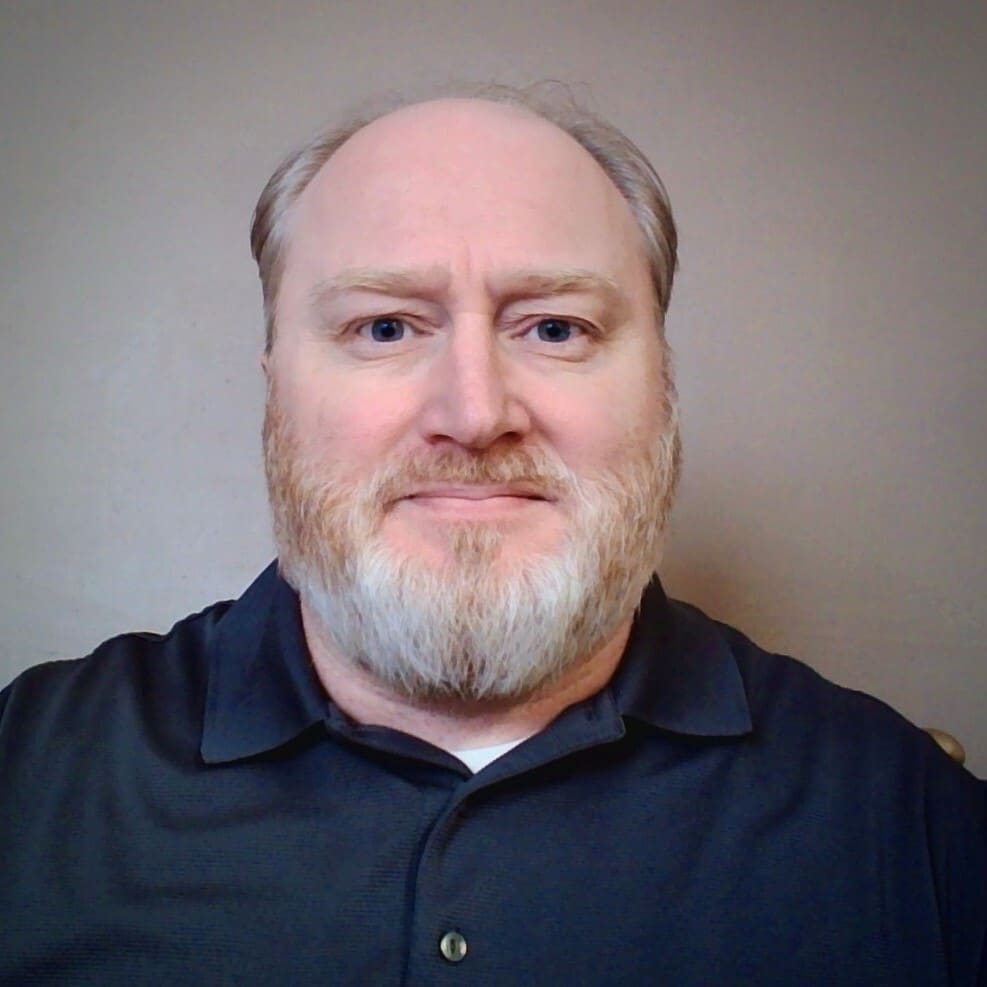 A person with a beard is wearing a dark polo shirt and looking at the camera. The background is plain and light-colored.