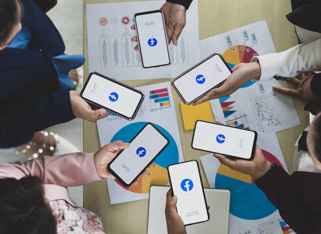 A group of people sits around a table, holding smartphones displaying the Facebook logo, with various documents and charts on the table.