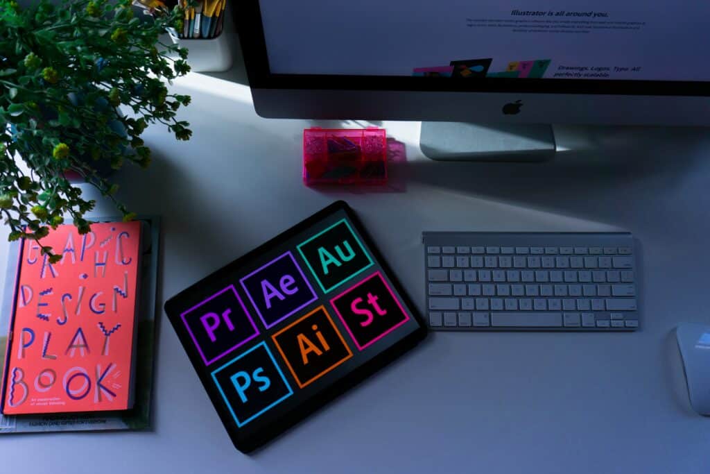 A modern workspace featuring an iMac, keyboard, and tablet displaying Adobe suite icons, a graphic design book, and a decorative plant and tape dispenser.