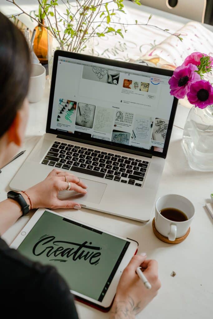 A person is working on a graphic tablet next to a laptop displaying design inspiration. A cup of coffee and fresh flowers are on the desk.