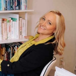 A person with shoulder-length blond hair is smiling at the camera, seated in front of a bookshelf filled with books. They wear a yellow scarf and black jacket.